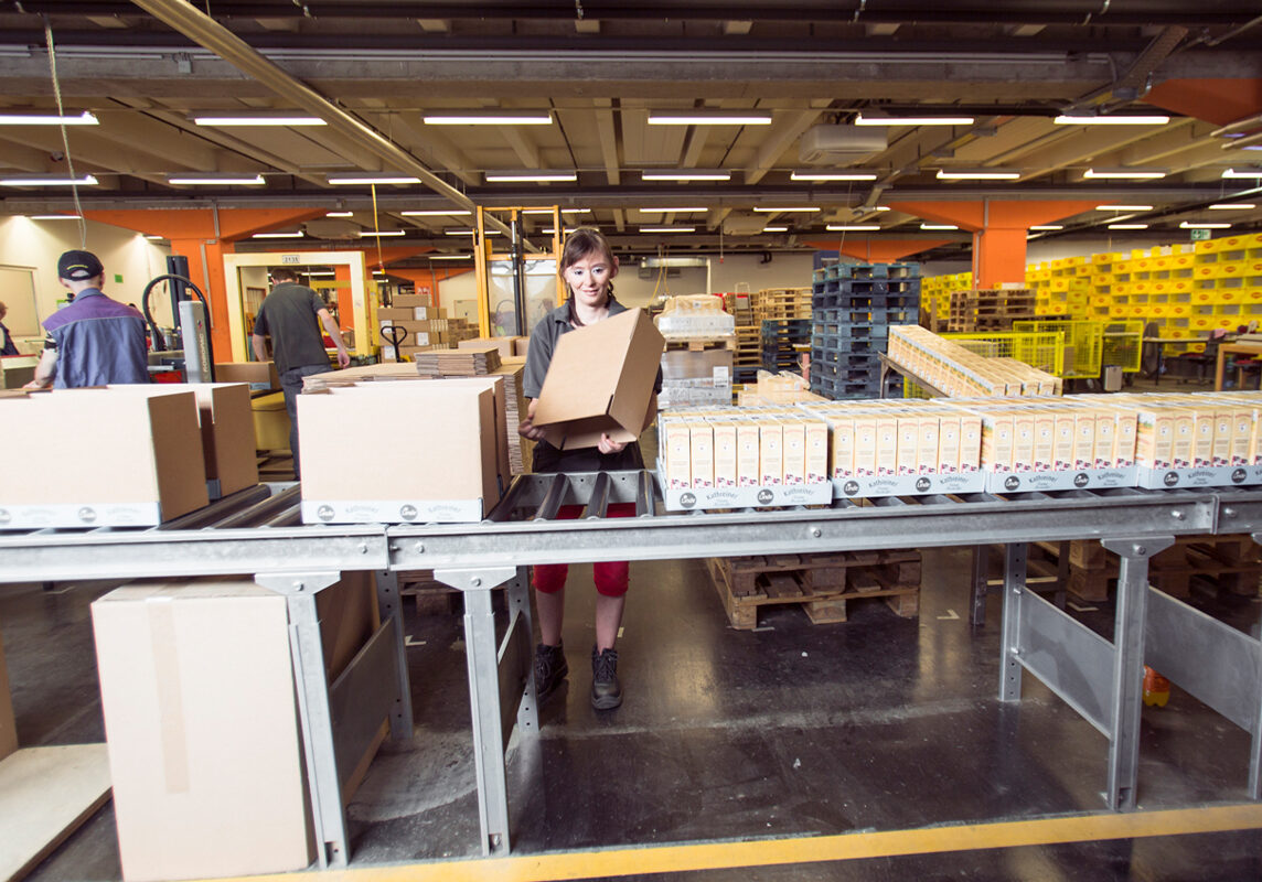 Eine FAB ProWork Mitarbeiterin steht am Laufband und verpackt Tray-Verpackungen.